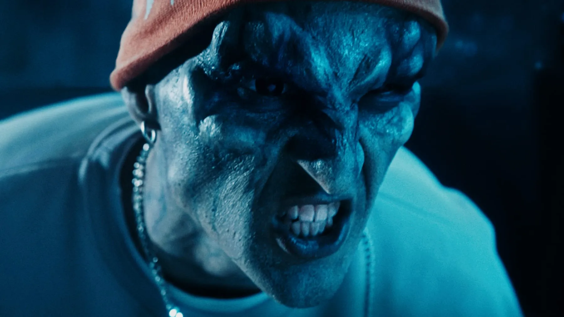 A player in alien prosthetic makeup, teeth bared under a red cap